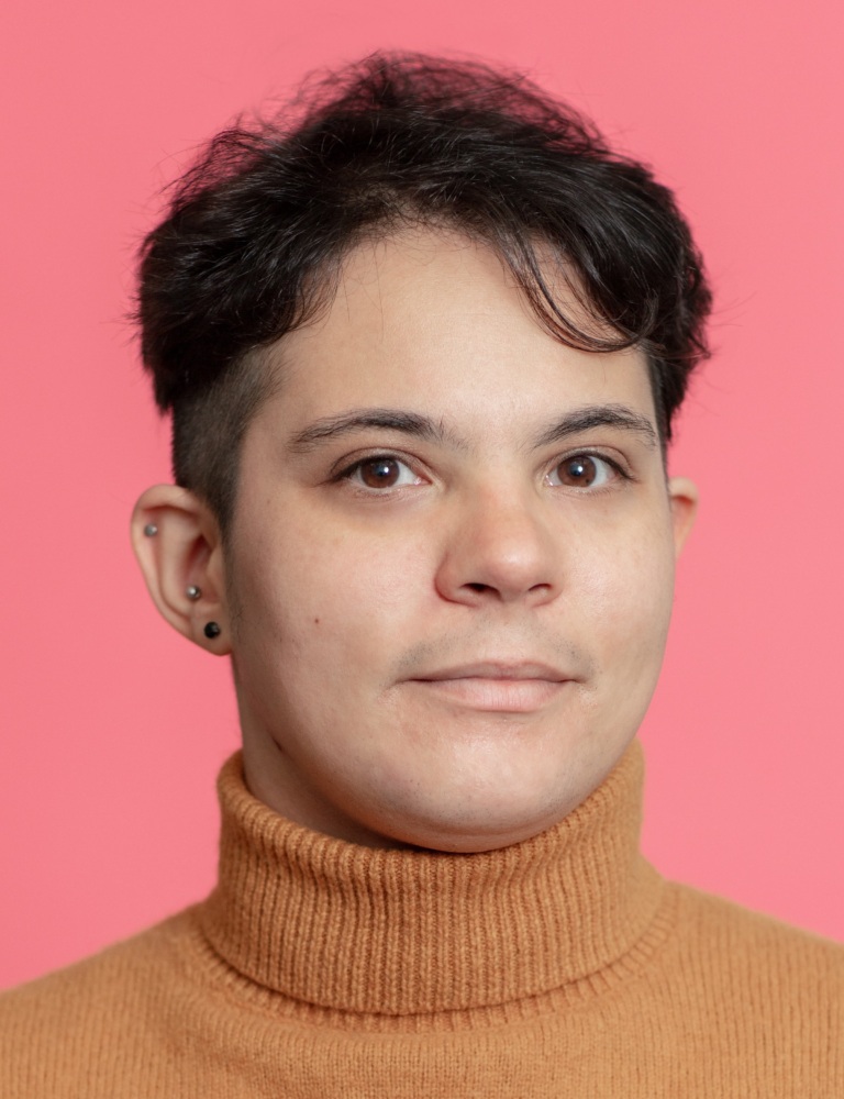 Person with short dark hair and a brown turtleneck sweater, looking directly at the camera against a pink background.